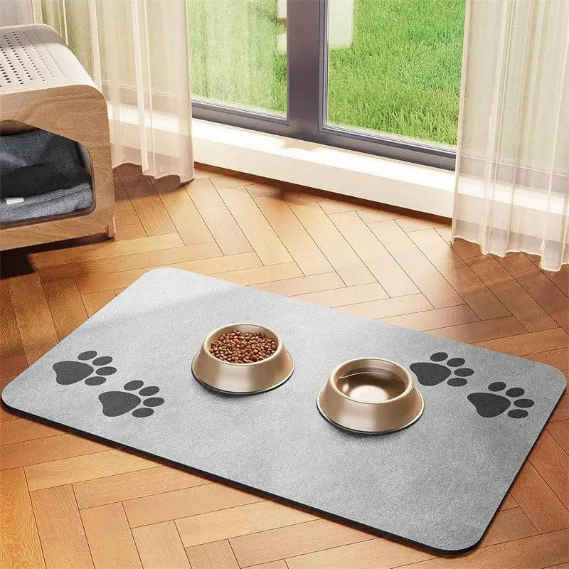 Gray suede-look absorbent pet feeding mat with a paw print at each end and two gold bowls, by a patio door.