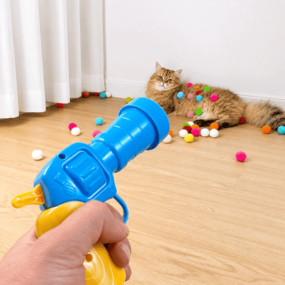 Hand firing the blue and yellow Noblias cat fetch toy ball launcher while a cat waits among colorful pom-pom balls