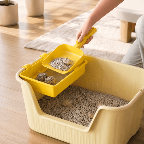 A hand lifting the yellow cat litter sifting tray out of a cream litter box with clumps caught in the mesh.