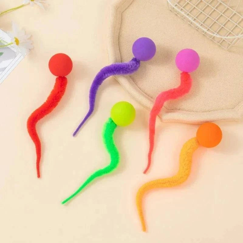 Five cat toy balls with fuzzy worm tails in red, purple, pink, green and orange on a beige backdrop