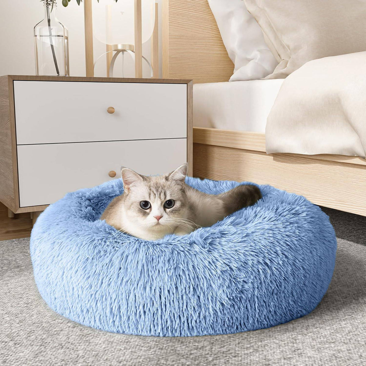 Light blue Noblias cloud calming donut bed with a cat settled inside beside a bed.