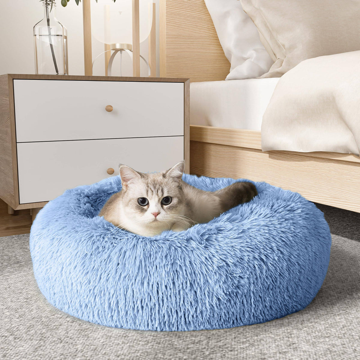 Cat resting in the light blue Noblias cloud calming donut bed beside a bedroom nightstand.