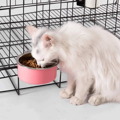 White long-haired cat eating dry food from the pink crate bowl clamped to a black wire cage