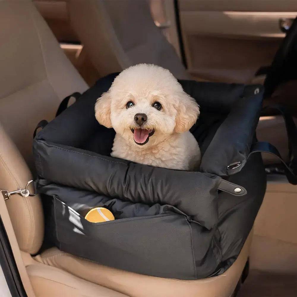 A small white dog sitting up in the black Noblias dog car seat bed inside a car.