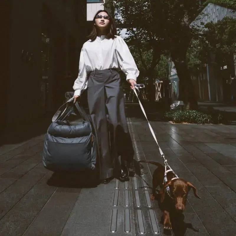 The gray Noblias dog car seat bed carried over the shoulder like a bag while walking a dachshund.