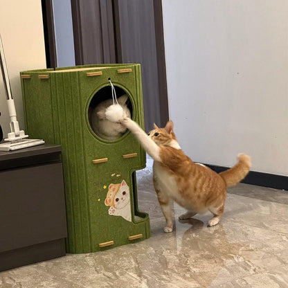 Ginger cat batting the hanging pompom on the Noblias felt cat house while a white cat looks out of the cave.