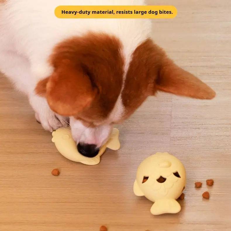 Dog chewing the light yellow fish treat dispenser toy on the floor with kibble scattered around, labeled heavy-duty material that resists large dog bites