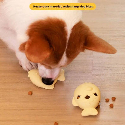 Dog chewing the light yellow fish treat dispenser toy on the floor with kibble scattered around, labeled heavy-duty material that resists large dog bites