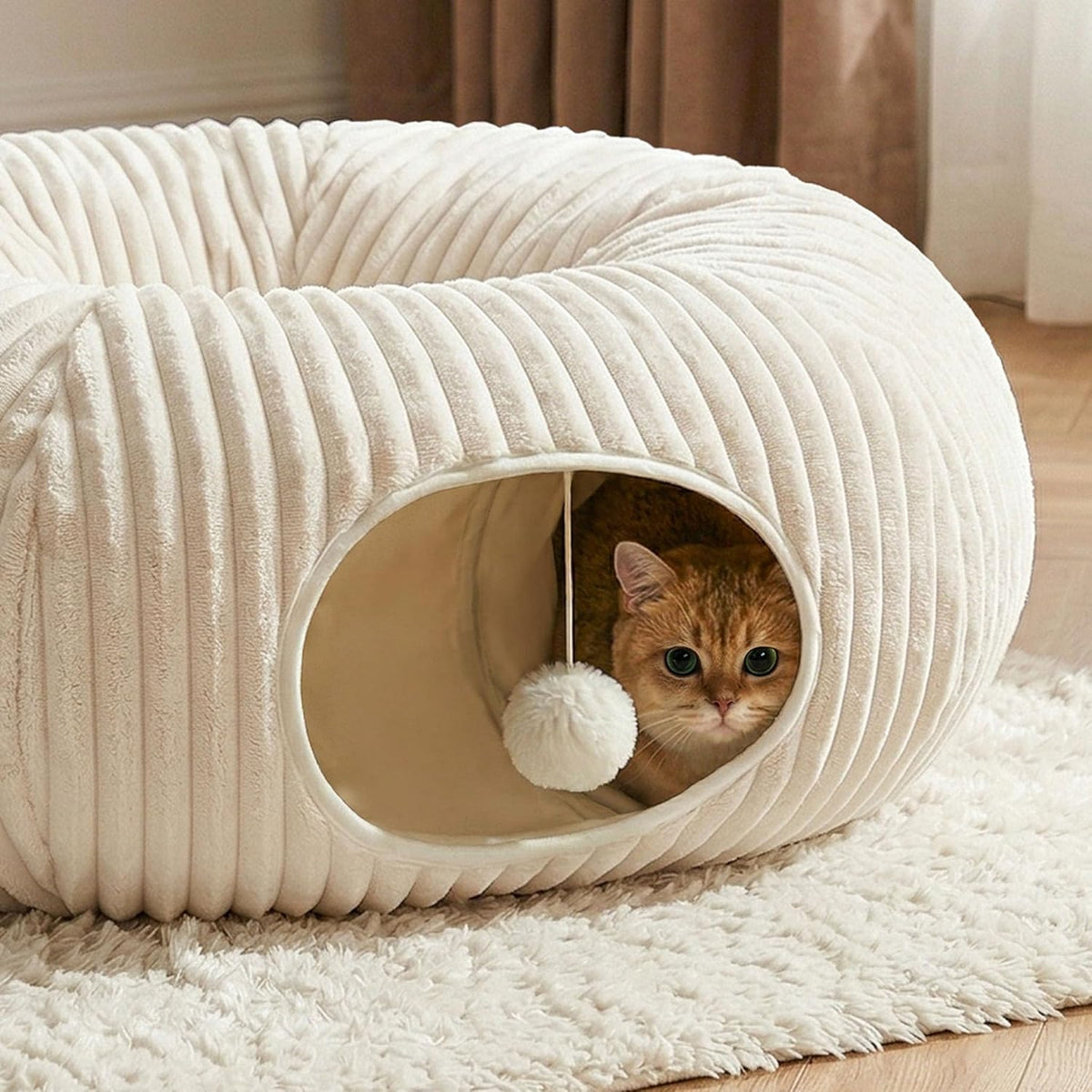 Ginger tabby cat sitting inside the tunnel opening looking out past the white pom-pom ball hanging at the entrance