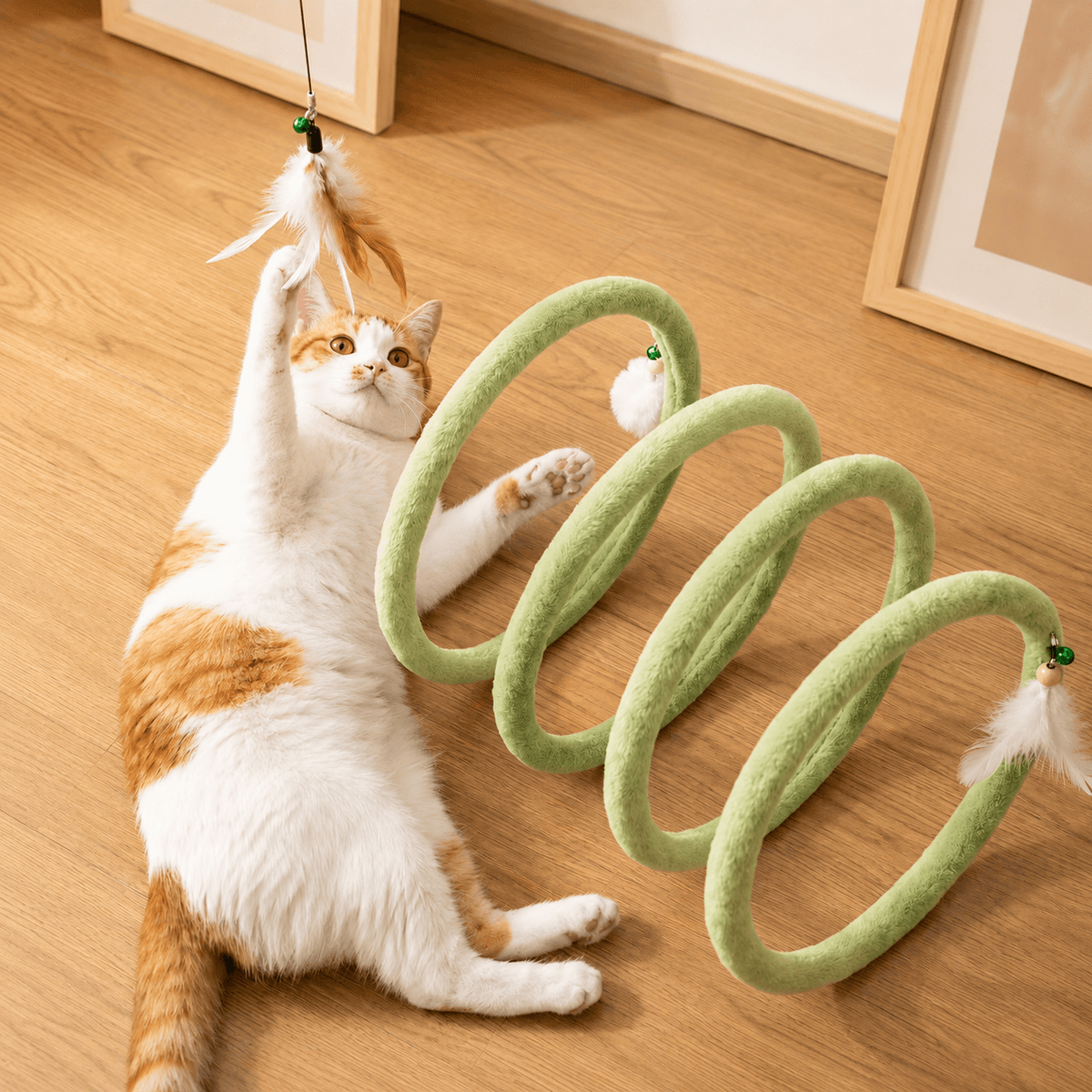 Cat lying on a wooden floor batting at the feather tip of the green spring cat tunnel