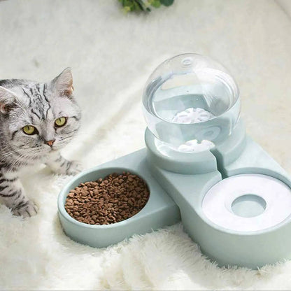 Gray cat lying beside the light blue Noblias gravity pet water dispenser with its globe reservoir and a bowl of kibble