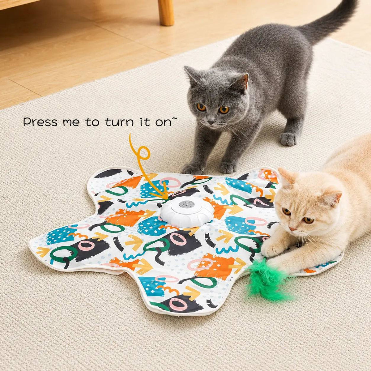 White hide and seek cat toy mat on a carpet with a gray cat and a cream kitten stalking it, labeled press me to turn it on