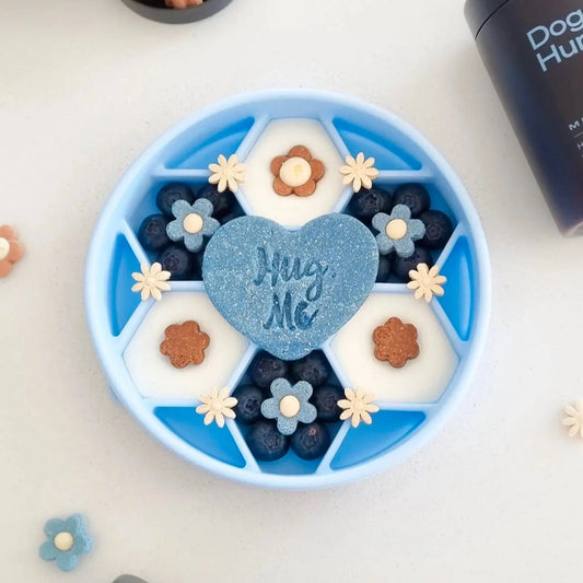 Blue Noblias honeycomb slow feeder dog bowl filled with blueberries, yogurt and flower shaped biscuits around a heart shaped treat.