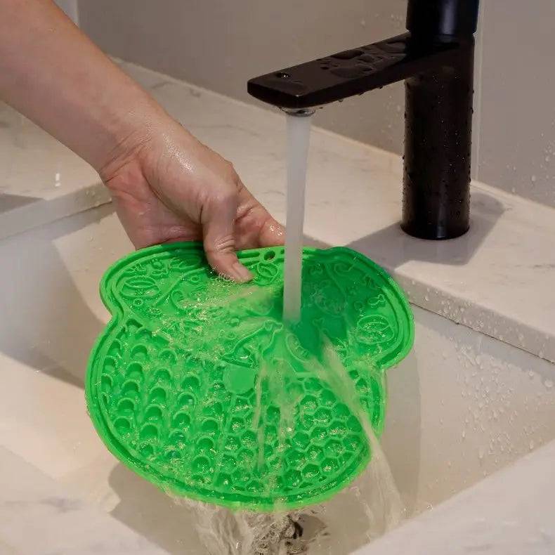 Green silicone lick mat being rinsed under a running kitchen tap.