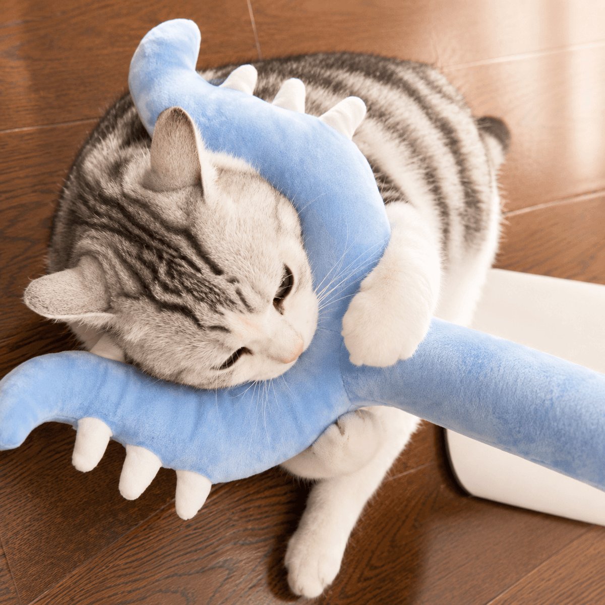 A silver tabby cat lying on a wooden floor with the blue plush head resting over its neck.