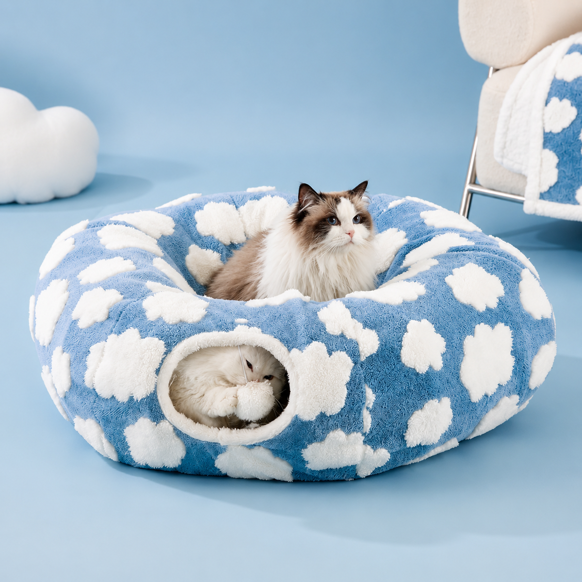 Blue plush cat tunnel bed with a white cloud pattern, one cat resting on the rim and another inside the tunnel opening