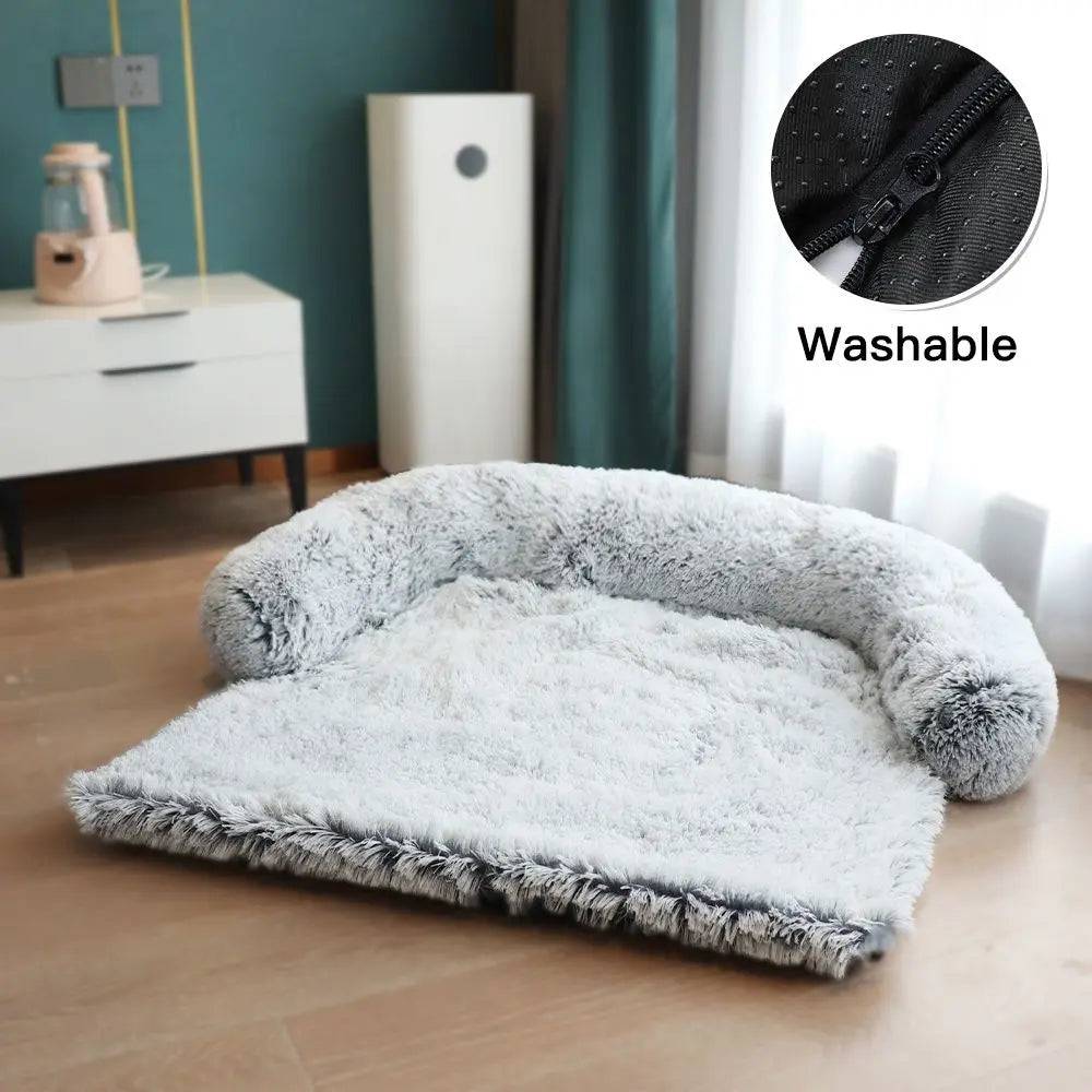 Light gray plush dog couch bed with its U-shaped bolster and washable zip cover, on a wood floor.