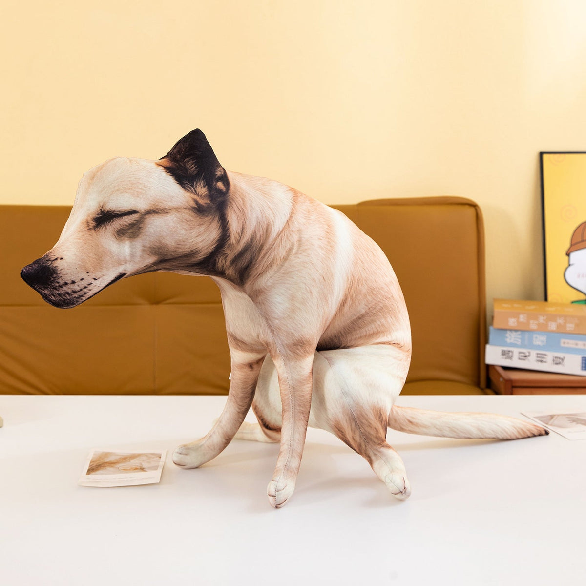 Funny pooping dog plush toy sitting on a table in front of a tan sofa, seen from the front.