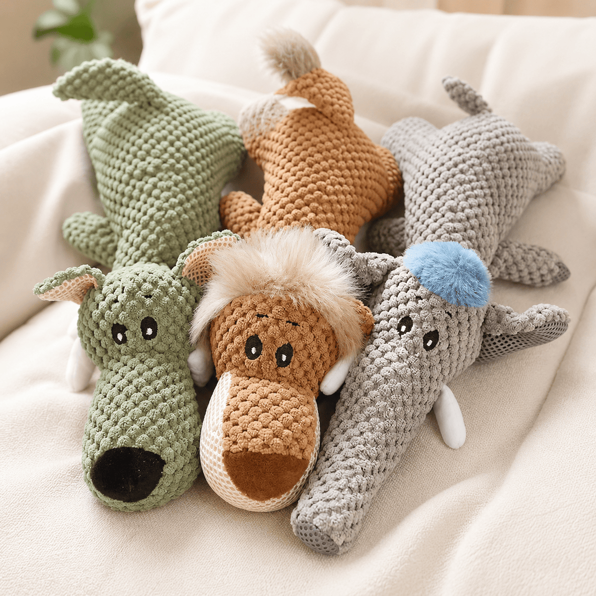 Green wolf, orange lion and gray elephant squeaky plush animal dog toys lying together on a sofa.