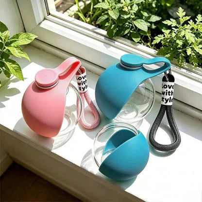 Pink and blue travel dog water bottles on a windowsill, the blue silicone sleeve flipped open to form the drinking cup.